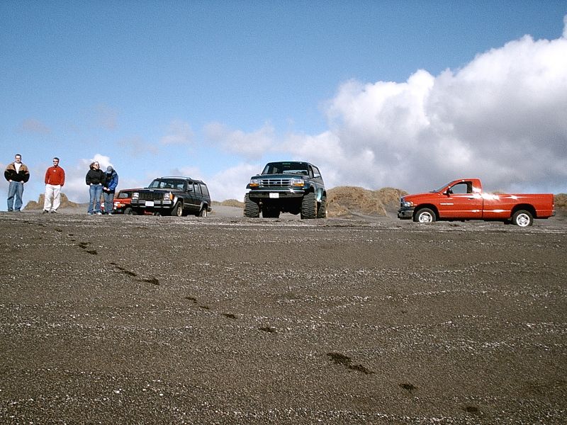 Lifted dark Bronco, dark SUV, and red pickup parked on gravel flat with people standing nearby