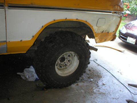 1978 Ford Bronco - The rear wheel well and large tire of a two-tone yellow and white vehicle are visible, with another car in the background.