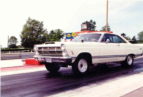 1967 Ford Fairlane - A white 1966 Ford Fairlane is captured mid-race on a drag strip, a prominent hood scoop and racing tires.