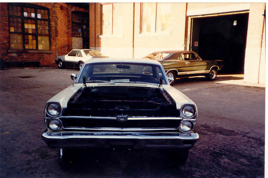 Front view of a cream Ford Fairlane with hood removed outside a brick shop