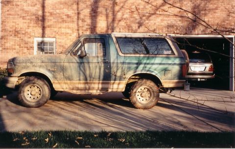 1996 Ford Bronco - A weathered 1996 Ford Bronco sits outside a garage, partially obscuring another vehicle within.