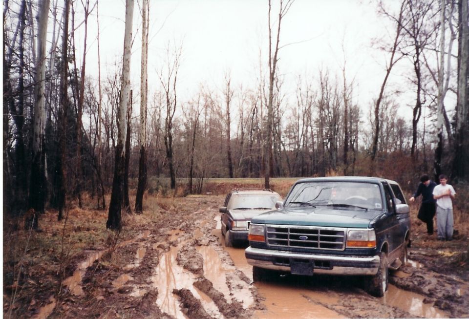 Dark green Ford Bronco stuck in a muddy woodland trail with another SUV and two onlookers behind