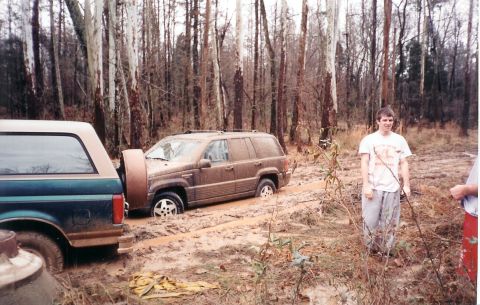 1996 Ford Bronco - Two off-road vehicles are stuck in deep mud in a wooded area, with a young man observing the scene.