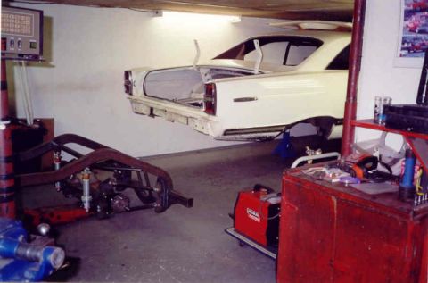 1967 Ford Fairlane - A white sedan is suspended in a garage, with its trunk open, as a separate chassis sits nearby.