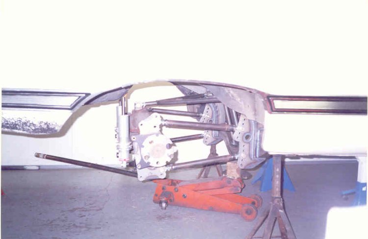 Rear underside of stripped car on jack stands showing tube frame, axle brackets and orange floor jack