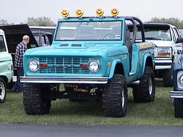 Turquoise lifted early Ford Bronco, front view, roll bar, big tires, smiley-face lights on windshield.