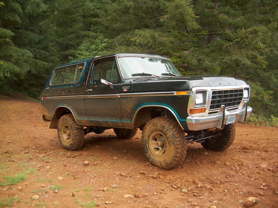 Mud-splattered dark green lifted Ford Bronco on big tires parked on red dirt in a forest clearing