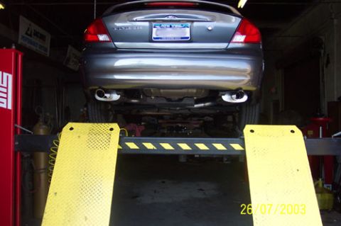 2003 Ford Taurus - A gray sedan is raised on a hydraulic lift in a service bay, showing its undercarriage and dual exhaust system.