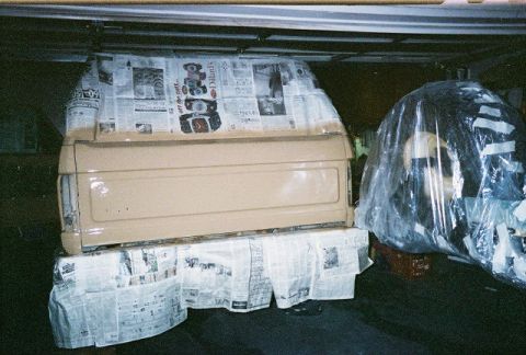 1983 Ford Bronco - A pickup truck bed is being prepped for paint, with newspaper covering the body panels and a tire wrapped in plastic nearby.