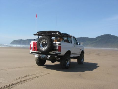 1995 Ford Bronco - A white Ford Bronco is parked on a sandy beach with the ocean and hills in the distance under a clear blue sky.