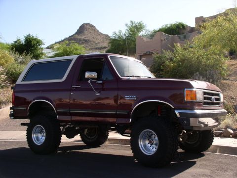 1990 Ford Bronco - This lifted Ford Bronco has a two-tone paint scheme and is parked on a street with a desert landscape in the background.