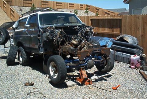 1986 Ford Bronco - A partially disassembled two-tone gray/silver SUV sits on gravel with its front end stripped, engine and wiring harness exposed and the fro…
