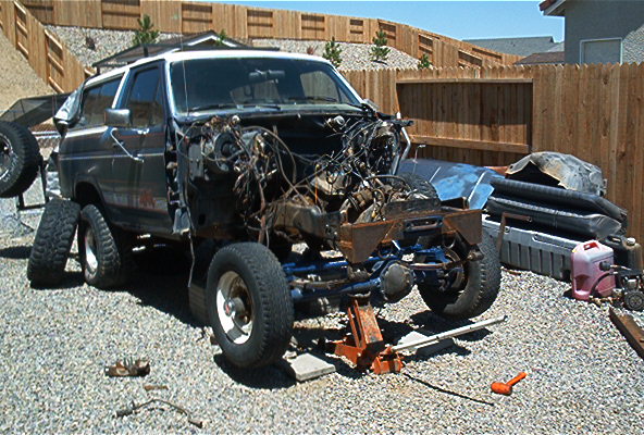 Gray SUV with front end torn down, exposed engine and wiring, tools on gravel by fence