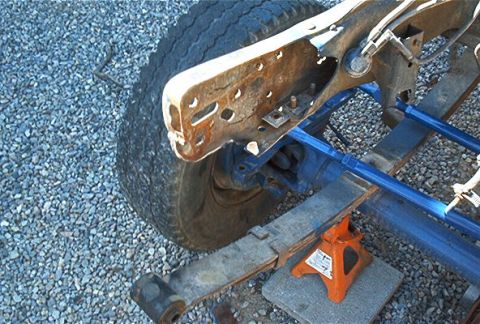 1986 Ford Bronco - Close-up of a rear suspension undergoing service, showing a rusty leaf spring and shackle mount, a blue axle/frame member, and the tire sup…