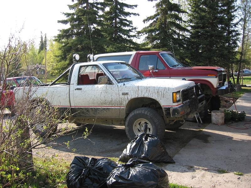 Muddy white S-10 4x4 pickup with roof light bar parked beside a red Ford Bronco in a gravel driveway