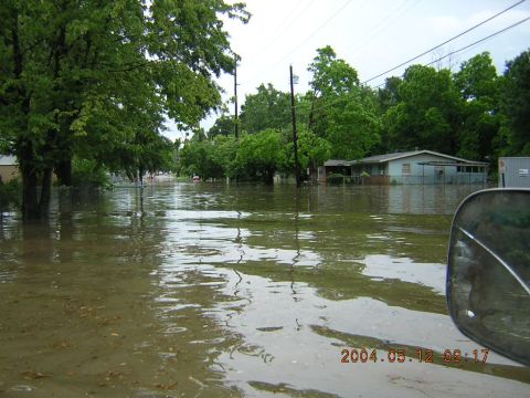 1984 Chevrolet Blazer - A street is submerged in floodwaters, with houses and trees visible on the sides, under an overcast sky.