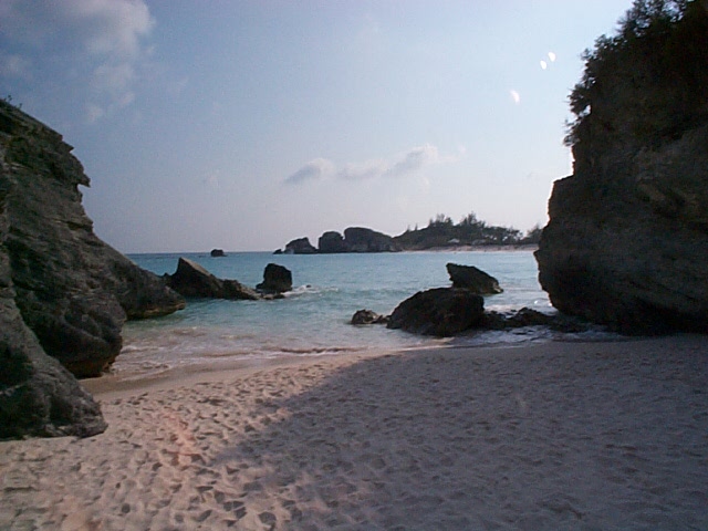 Sandy beach with large rocks and calm blue ocean under a partly cloudy sky. Watermark reads SUPERMOTORS