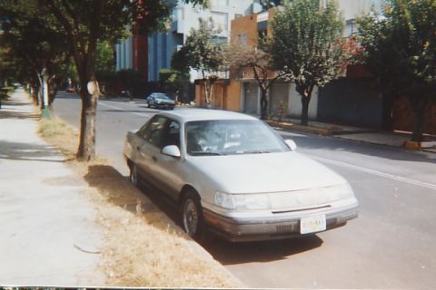 1991 Ford Taurus - A silver sedan is parked on the side of a street lined with trees and buildings. Another car is visible in the distance.