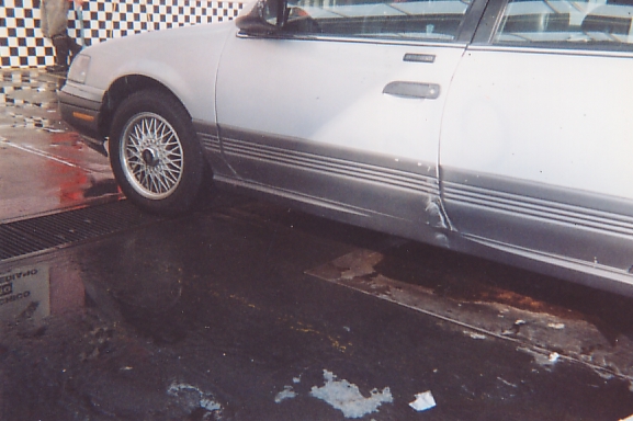 Silver sedan side view showing front wire wheel and dented rear door on wet pavement.