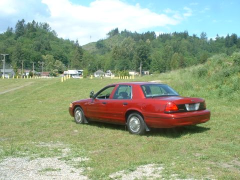 1999 Ford Crown Victoria - A red sedan sits in a grassy field with trees and hills in the background.