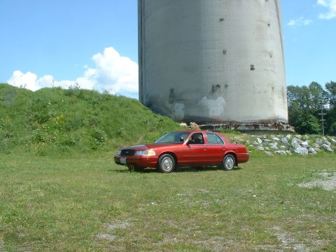 1999 Ford Crown Victoria - A red 1999 Ford Crown Victoria is parked on a grassy field next to a large concrete silo.