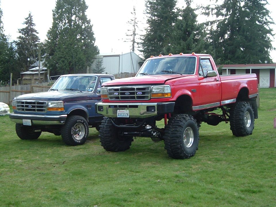 Blue Ford Bronco and lifted red Ford F-250 pickup on mud tires parked on grass