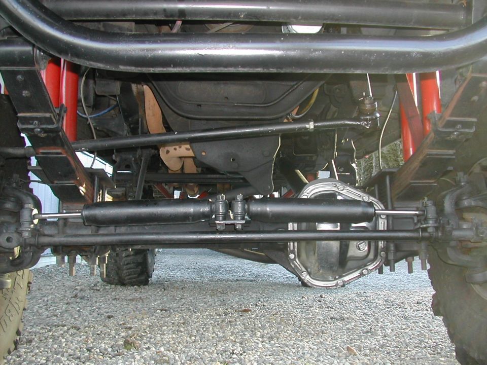 Under-truck view of solid front axle, dual steering stabilizers, and leaf springs on a lifted 4x4 over gravel