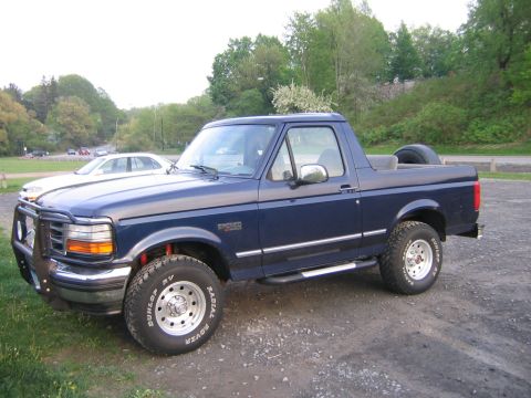 1994 Ford Bronco - A blue Ford Bronco is parked on a gravel lot with trees in the background.