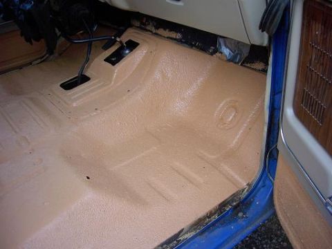 1980 Ford Bronco - The tan textured floor of a blue vehicle interior is visible, with pedals and a portion of the door panel.