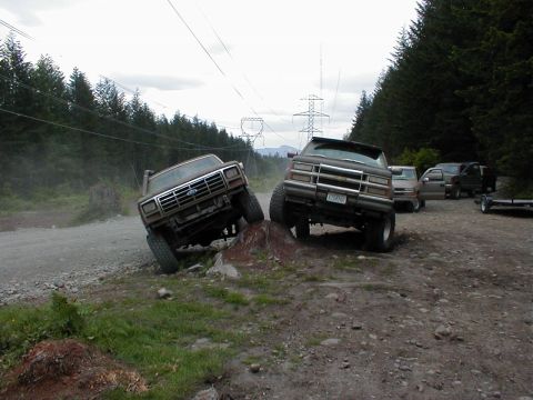 1983 Ford Bronco - Two mud-covered lifted 4x4s flexing over a dirt mound on a gravel staging area, an older Ford truck on the left with one front tire climbin…
