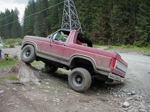 1983 Ford Bronco - A red SUV navigates a rocky off-road trail, with a power line tower in the background.