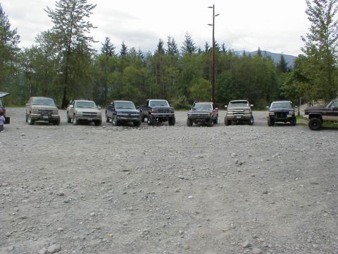 1983 Ford Bronco - Multiple pickup trucks and SUVs are parked in a line on a gravel lot with trees in the background.