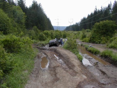 1983 Ford Bronco - A dark 4x4 truck tipped at a steep angle in a deeply rutted mud trail with several spectators standing alongside it, seen from down-trail.…