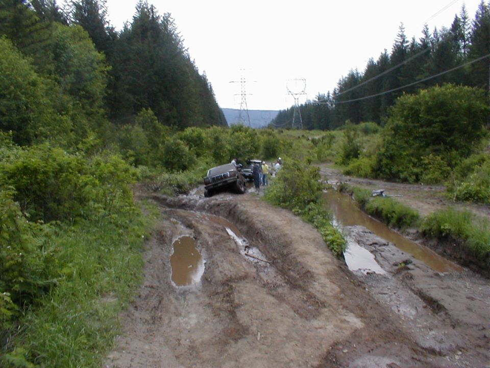 Dark 4x4 truck leaning in deep mud ruts on a powerline trail with onlookers