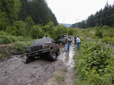 1983 Ford Bronco - Several people watch as a lifted Ford truck struggles through a muddy off-road trail.