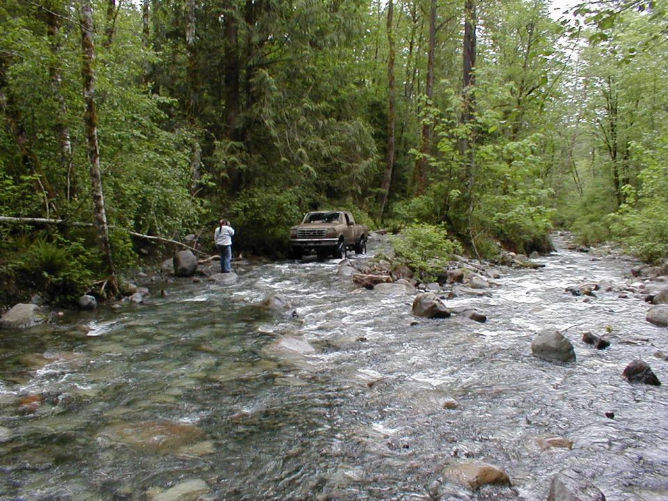 Brown pickup truck crossing stream, forest setting