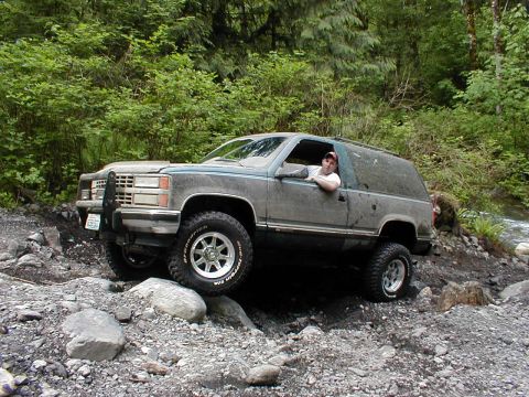 1983 Ford Bronco - SUV driving off-road on a rocky path with trees in the background.