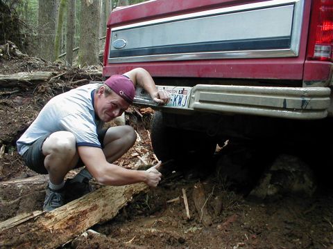 1983 Ford Bronco - A man inspects the rear bumper of a red pickup truck that appears to be stuck in the mud on a wooded trail.