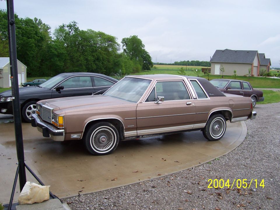 Brown two-door Ford LTD Crown Victoria with wire wheel covers on a wet rural driveway