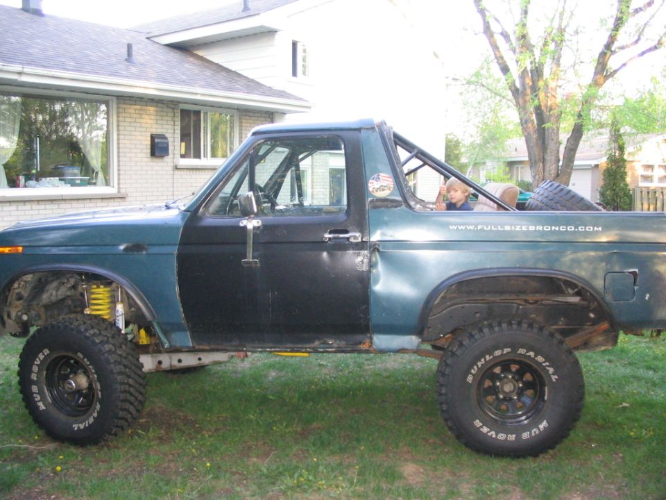 Side view of a modified Ford Bronco with a roll cage and mismatched doors. A child is visible in the passenger seat