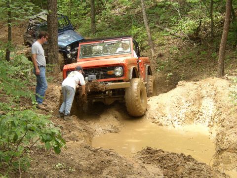 1995 Ford Bronco - An orange first-generation Ford Bronco navigates a muddy trail, with a blue Jeep visible behind it.