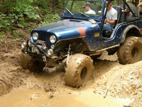 1995 Ford Bronco - A blue Jeep navigates a muddy trail with large, aggressive tires kicking up spray.
