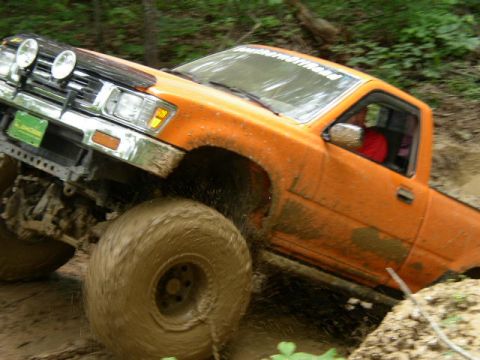 1995 Ford Bronco - An orange pickup truck navigates a muddy off-road trail, its front-left tire deep in the mire.