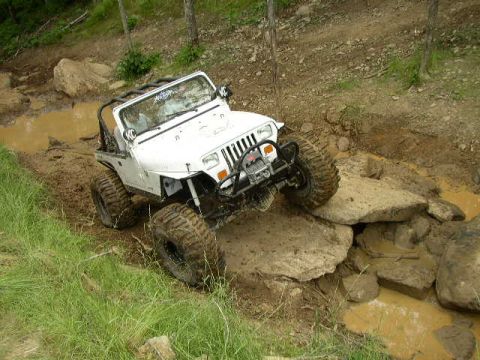 1995 Ford Bronco - A white Jeep Wrangler tackles a challenging off-road course with large rocks and mud pits.