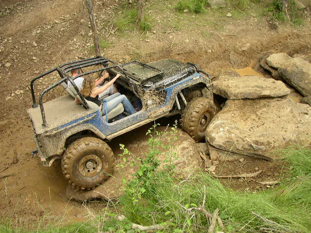 Mud-covered blue Jeep Wrangler navigating large rocks on an off-road trail