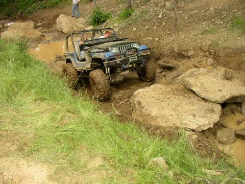 1995 Ford Bronco - A blue Jeep Wrangler tackles a challenging off-road course, splashing through mud and climbing over large rocks.