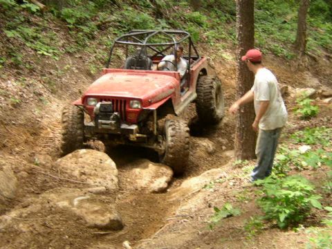 1995 Ford Bronco - A red off-road vehicle tackles a challenging rocky terrain on a wooded trail.