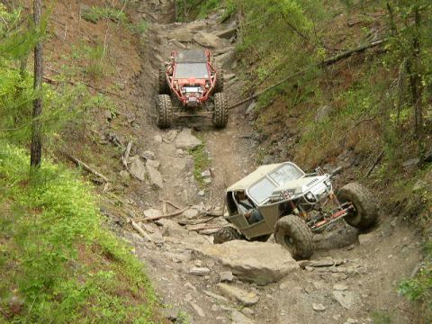 1995 Ford Bronco - Two custom off-road vehicles navigate a steep, rocky trail through a wooded area.