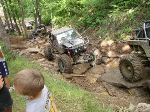 1995 Ford Bronco - Multiple off-road vehicles traverse a challenging, muddy, and rocky terrain in a wooded area.