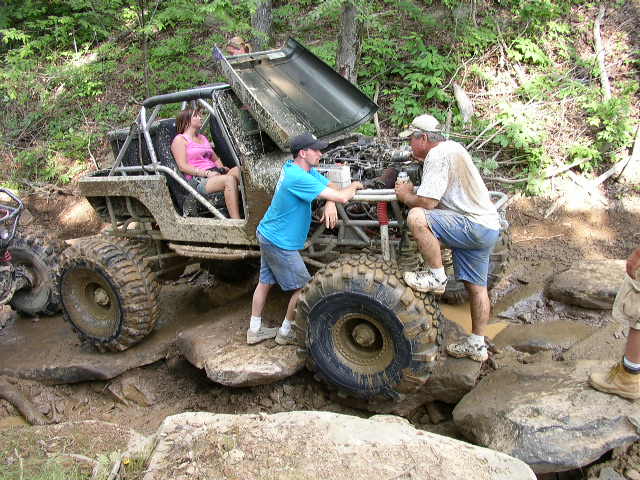 Mud-covered off-road vehicle with open hood, people around it, on rocky terrain